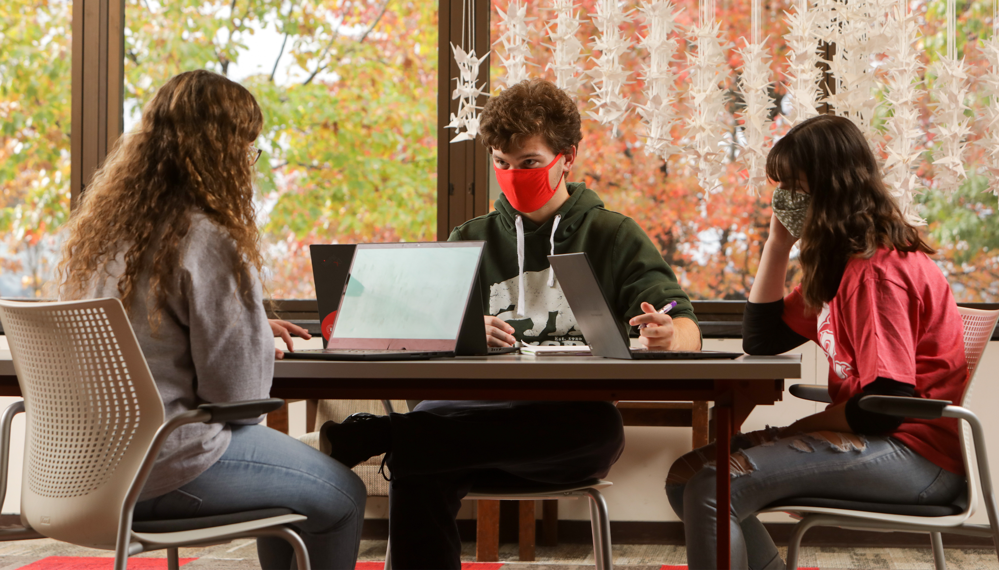 Three students wearing face masks, sitting at a table with each other using their laptops.
